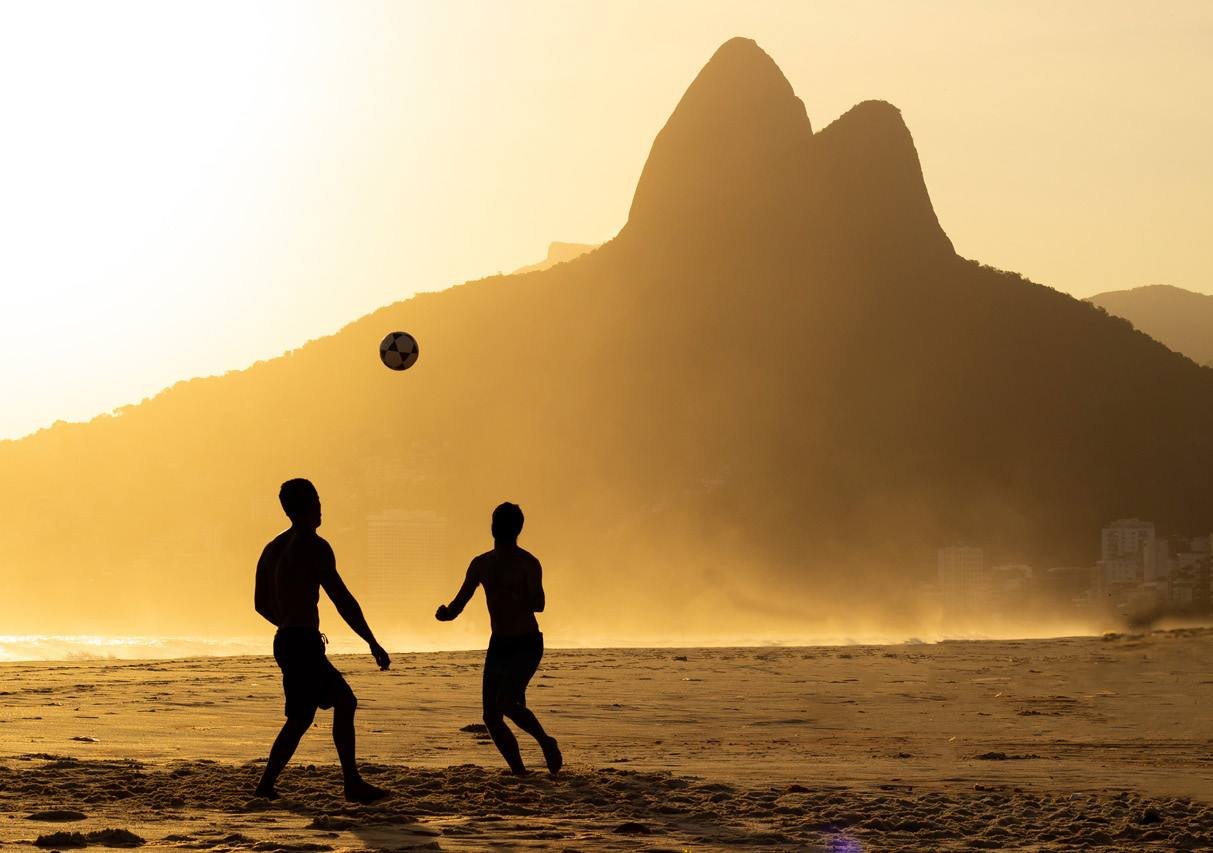 Silhuetas jogando futebol na praia de Ipanema ao pôr do sol, com o Morro Dois Irmãos